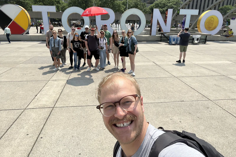 Grupa osób pozujących przed dużymi listami „Toronto” na placu, z jedną osobą na pierwszym planie robiącą sobie selfie.