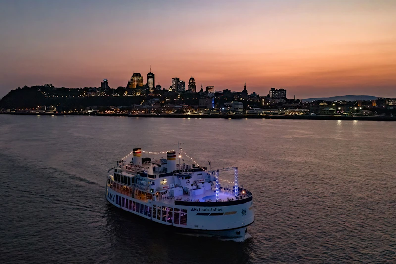 Rozświetlony prom płynący po rzece o zmierzchu, z panoramą miasta z oświetlonymi budynkami w tle.