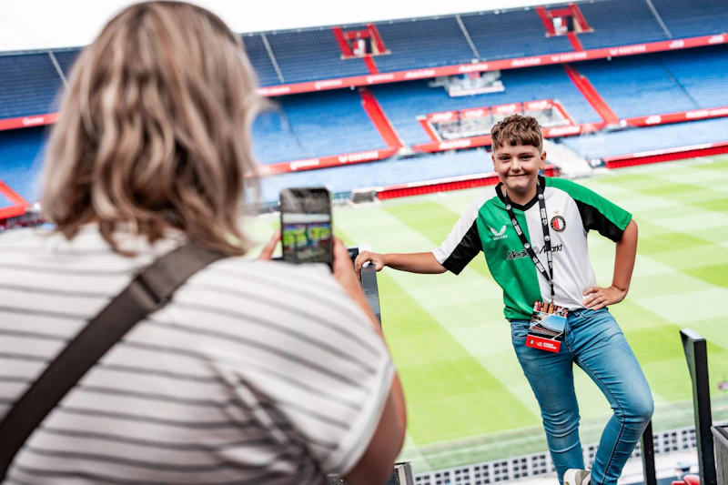 Chłopiec w zielono-białej koszulce pozuje do zdjęcia na stadionie, a osoba rejestruje ten moment w telefonie.