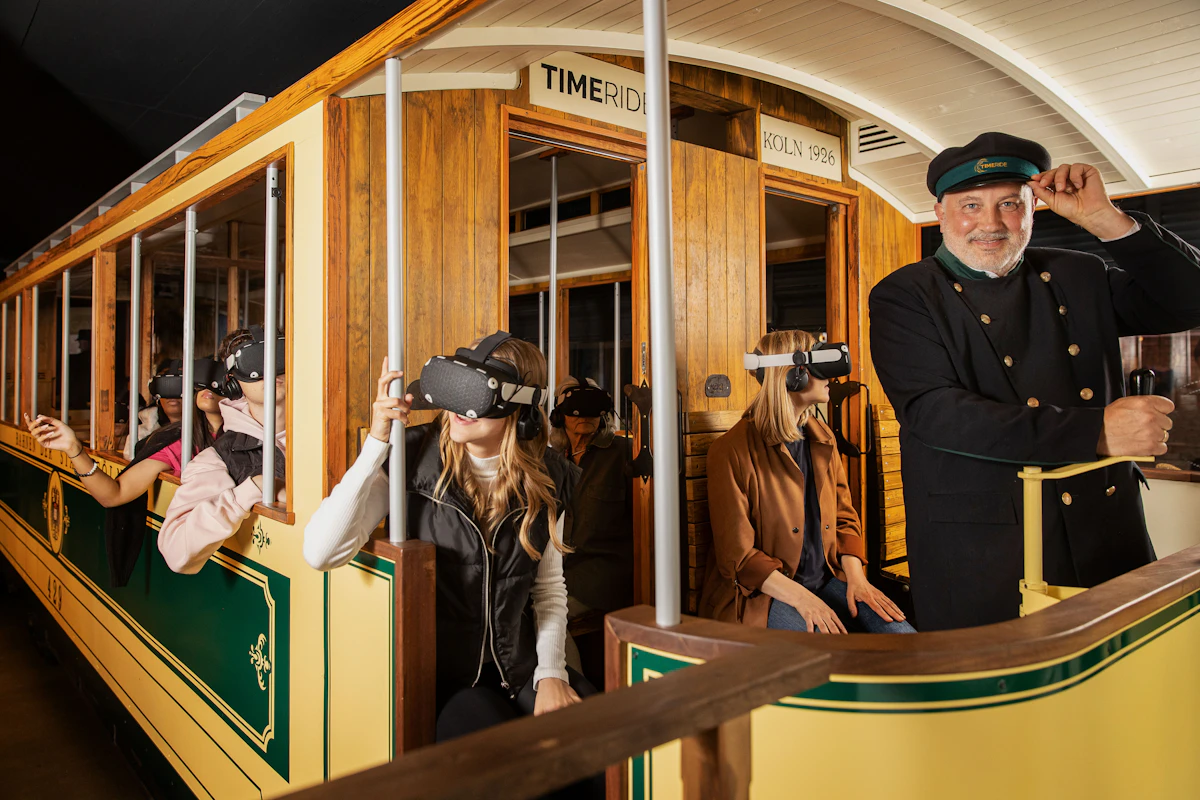 Dyrygent i trzy osoby w goglach VR wewnątrz drewnianego zabytkowego tramwaju z szyldem „Köln 1926”.