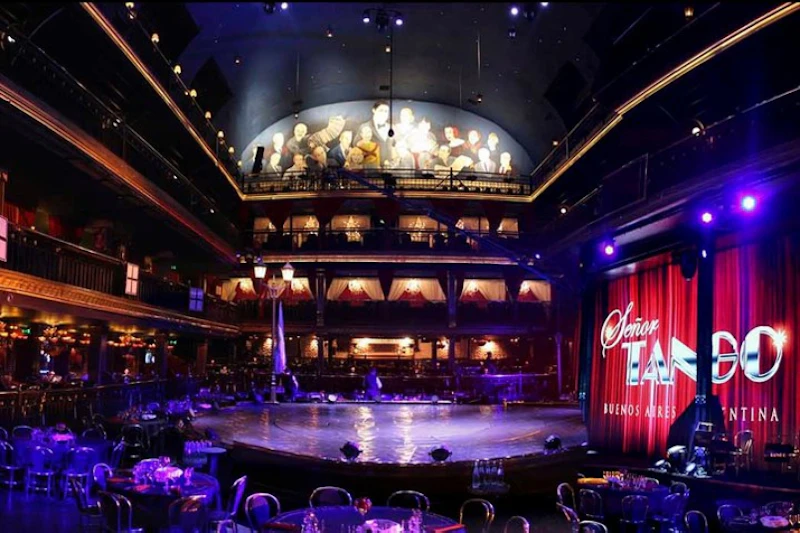 Sala koncertowa z pustymi stolikami, sceną, balkonami i malowanym muralem na suficie. Czerwone zasłony z tekstem „Señor Tango Buenos Aires”.