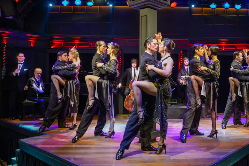 Pary wykonują tango na scenie w eleganckim stroju, z zespołem grającym w tle. Scena oświetlona jest żywymi kolorami.