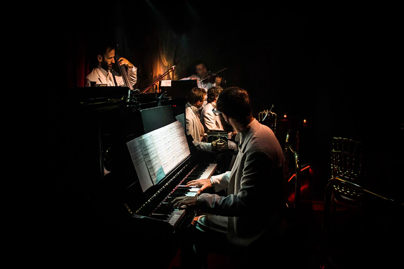 Na scenie występuje zespół jazzowy w garniturach, na pierwszym planie pianista, a w tle basista, saksofonista i trębacz.