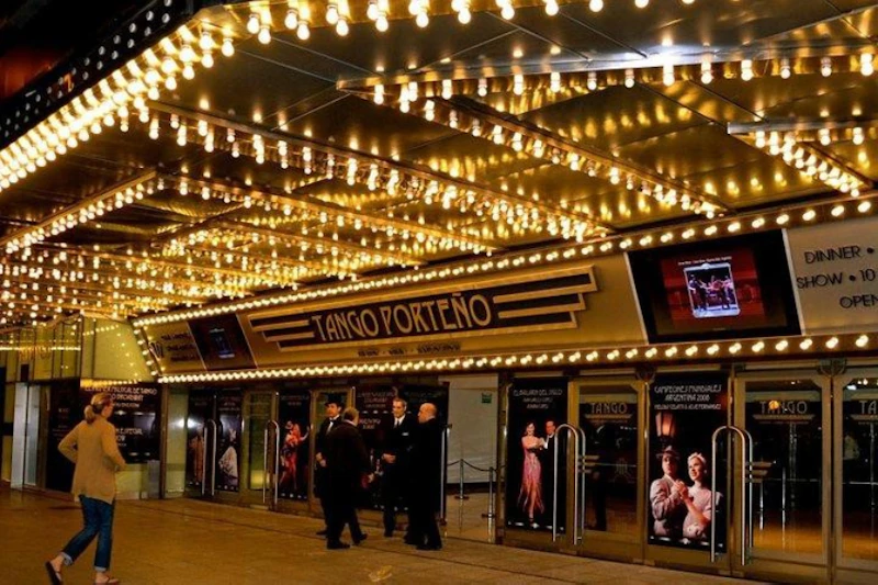 Podświetlane wejście do teatru z oznakowaniem „Tango Porteño”, plakaty tancerzy i osób w strojach formalnych stojących na zewnątrz.