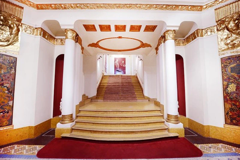 Eleganckie schody z białymi i złotymi kolumnami, czerwonym dywanem i ozdobnym sufitem prowadzące do portretu na szczycie.