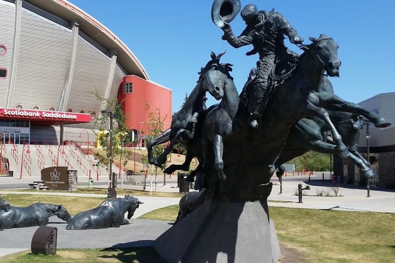 Posąg kowboja jadącego na bucking bronco, znajdujący się w pobliżu Scotiabank Saddledome na trawiastym placu z dodatkowymi posągami zwierząt.