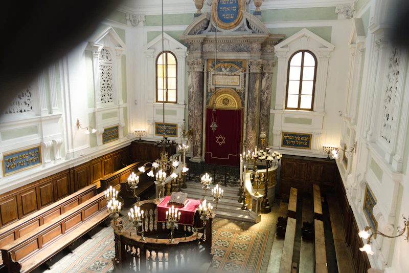 Ozdobne wnętrze synagogi z żyrandolami, drewnianymi ławkami, czerwoną kurtyną Tory i hebrajskimi napisami na ścianach.