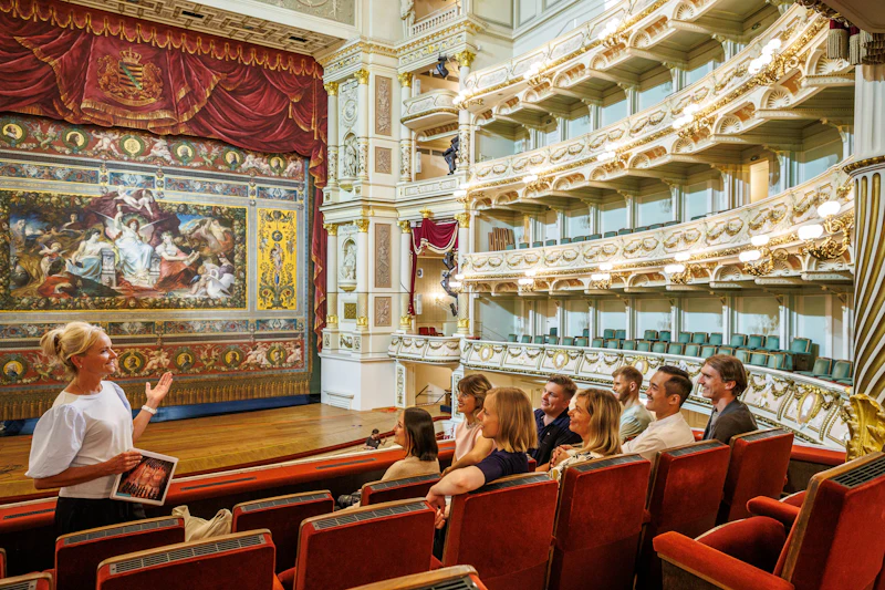 Grupa osób siedzących w pudełku teatralnym z ozdobnym wystrojem wnętrz, w tym czerwonymi siedzeniami i złotymi ozdobami.