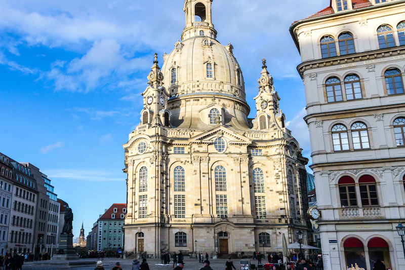 Wielki budynek w stylu barokowym z dużą kopułą w słoneczny dzień, ludzie spacerujący po pierwszym planie, otoczeni innymi budynkami.